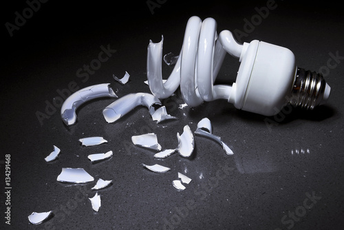 A Broken Compact Fluorescent CFL bulb lays shattered in shards of glass on a hard surface with a dark background