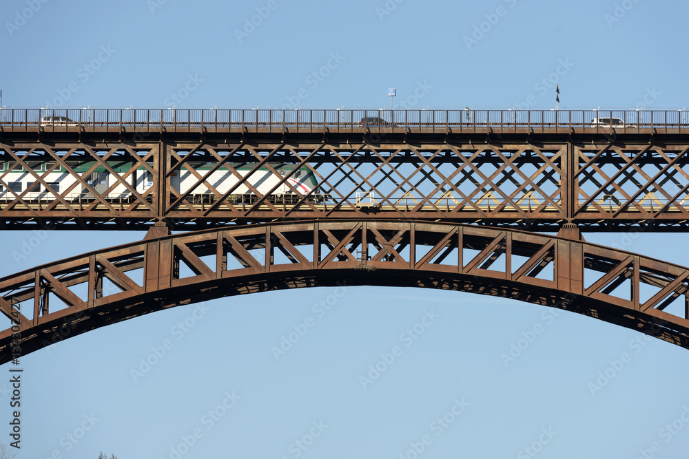 Naklejka premium train passes under cars on old iron bridge on river Adda , shot in bright light at Paderno, Lombardy, Italy.