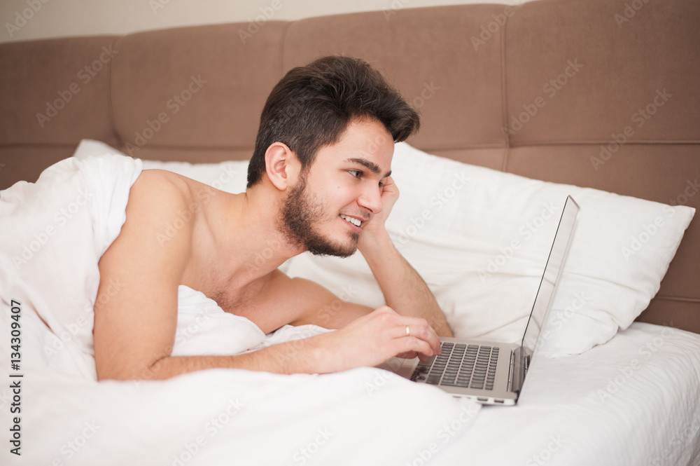 Young smiling man using laptop in bed at home Young smiling man using laptop in bed at home