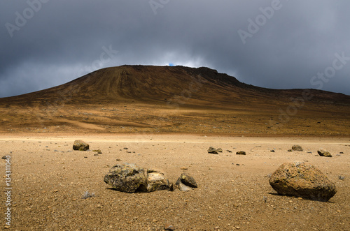 Desolate moonscape in the national park of Mount Kilimanjaro. Rocks lay a moon like landscape extending to a hill that is surrounded by dark clouds. 