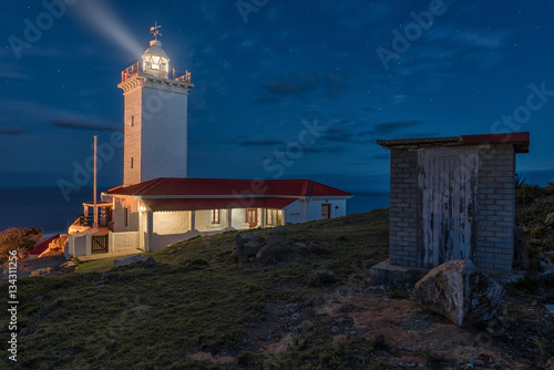 Lighthouse with rotating beacon and its light beam under blue cloudy night sky full of stars also showing a little shabby shed in the foreground