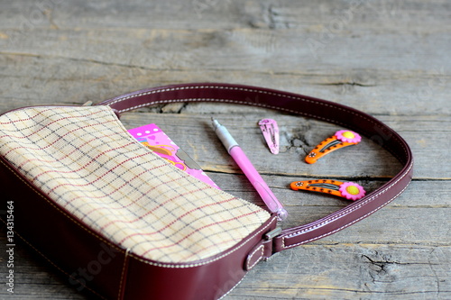 Children bag and accessories on wooden background. Small bag, notebook, pink pen, bright hair clips for girl. Children purse content. Closeup