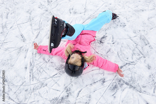 Child girl falling down on ice in snowy park during winter holidays. Wearing safety helmet. Winter children activities.