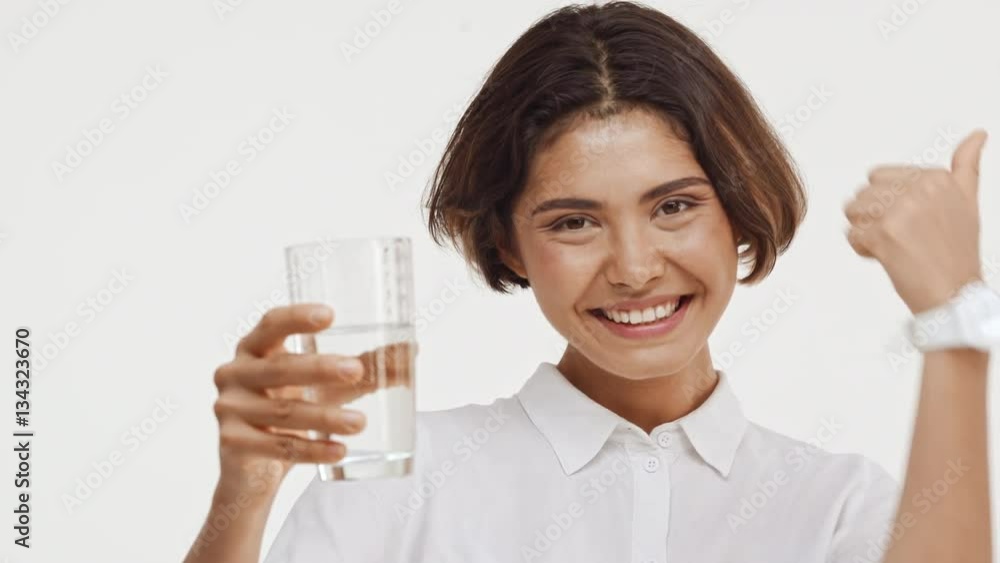 Young beautiful brunette east asian female in shirt showing glass with water smiling approving and drinking on white background Young beautiful brunette east asian female in shirt showing glass with water smiling approving and drinking on white background