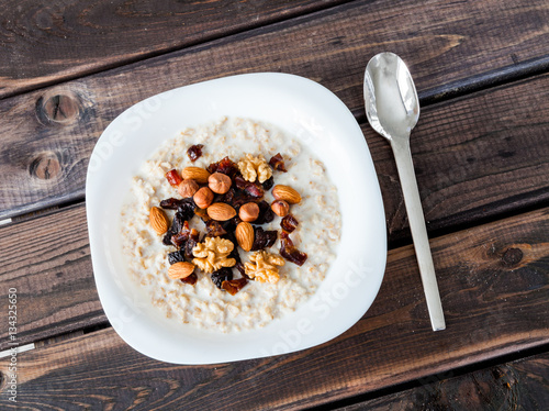 Bowl of homemade oatmeal porridge with dried plums, almonds, hazelnuts, walnuts and dates on old dark grey rustic wooden table with spoon. Hot and healthy food for Breakfast. 