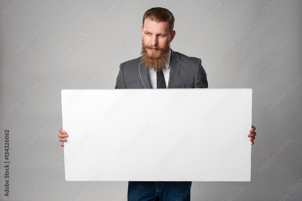 Hipster business man with beard and mustashes in suit standing holding white banner, looking away with thoughts, over grey background Hipster business man with beard and mustashes in suit standing holding white banner, looking away with thoughts, over grey background