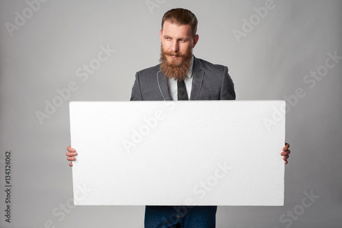 Hipster business man with beard and mustashes in suit standing holding white banner, looking away with thoughts, over grey background photo