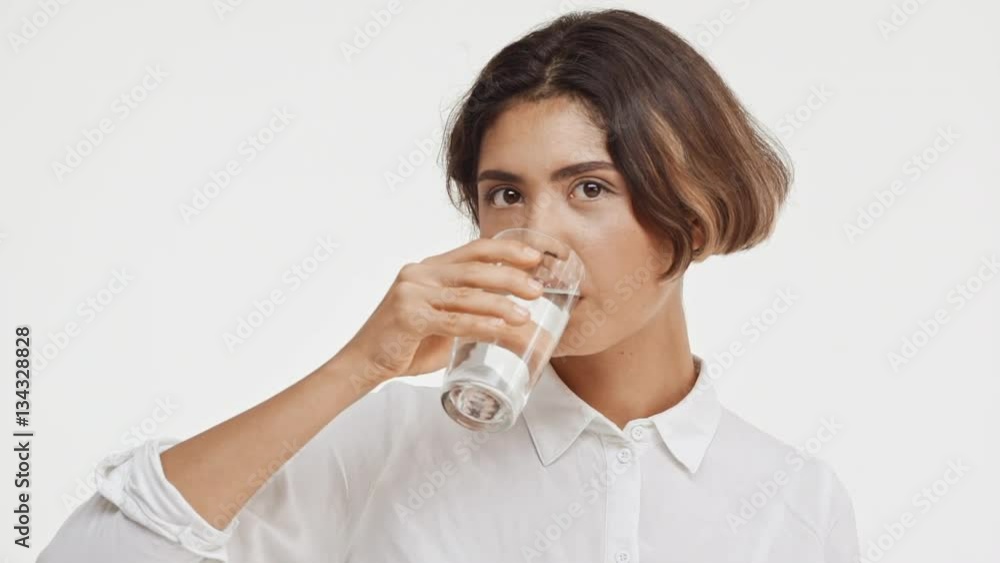 Young beautiful brunette east asian female in shirt drinking water from glass on white background in slowmotion Young beautiful brunette east asian female in shirt drinking water from glass on white background in slowmotion