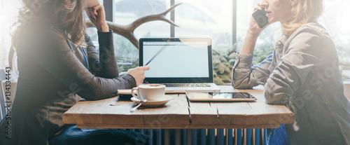 Teamwork, two young businesswomen sitting across table from each other. On table laptop, coffee cup and tablet computer. First girl showing pencil on computer screen, other is talking on cell phone.
