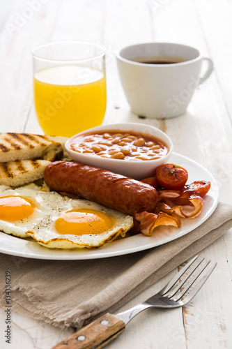 Traditional full English breakfast with fried eggs, sausages, beans, mushrooms, grilled tomatoes and bacon on white wooden background
