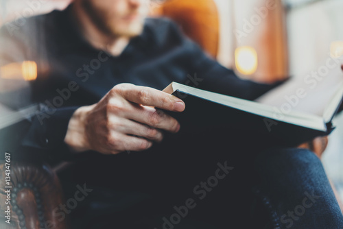 Closeup view of male hands holding book.Young bearded man relaxing at home while sitting in vintage chair.Selective focus on hand,blurred background.Horizontal, film effect.