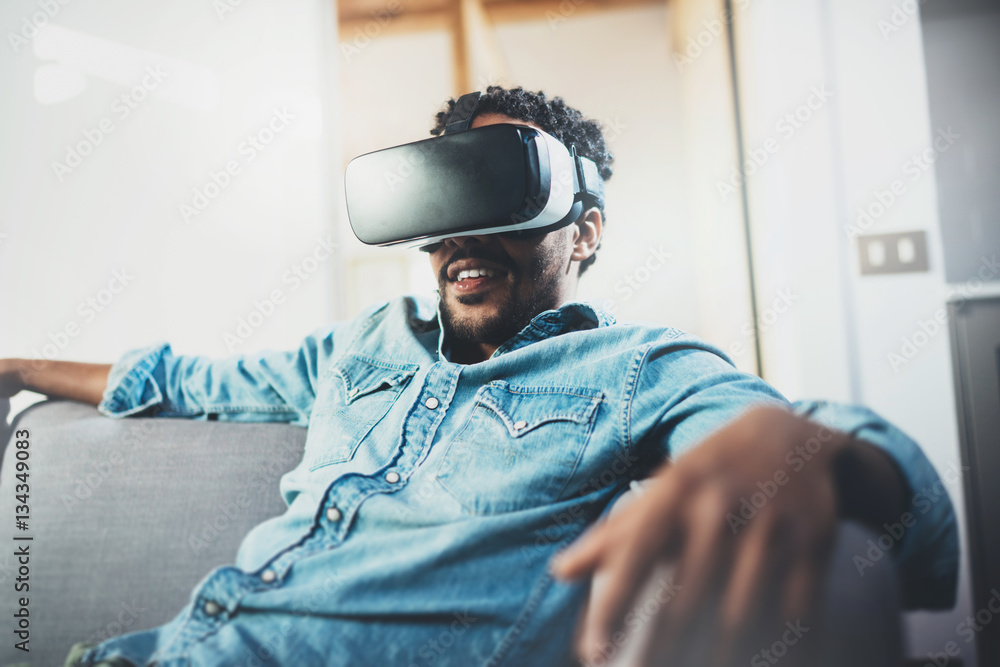 Smiling bearded african man enjoying virtual reality glasses headset or 3d spectacles while relaxing on sofa at modern home.Blurred background. Smiling bearded african man enjoying virtual reality glasses headset or 3d spectacles while relaxing on sofa at modern home.Blurred background.
