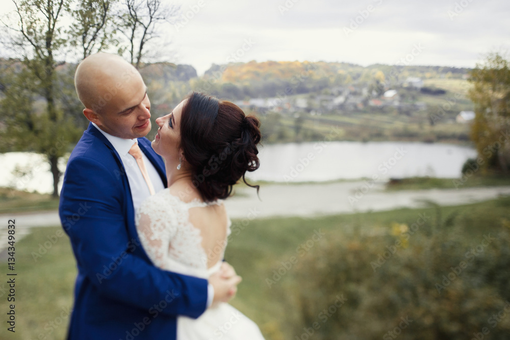 Groom admires bride hugging her on hill with great lake behind t Groom admires bride hugging her on hill with great lake behind t