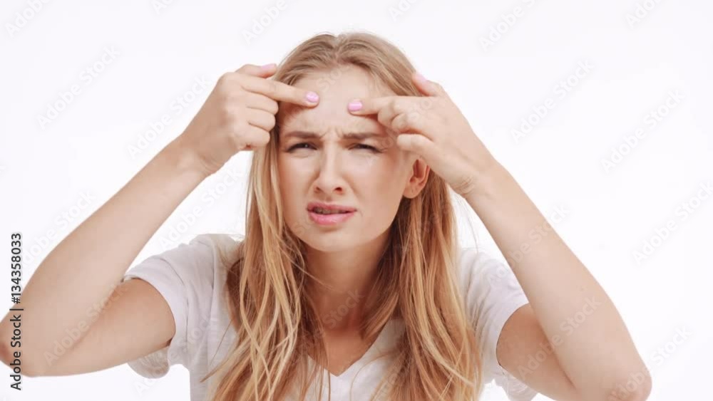 Young beautiful Caucasian blonde girl trying to find pimple on forehead on white background Young beautiful Caucasian blonde girl trying to find pimple on forehead on white background
