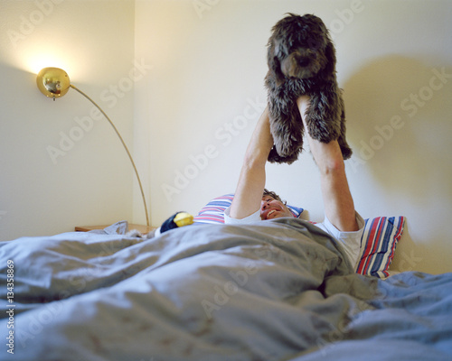 Man lying in bed holding dog in the air 