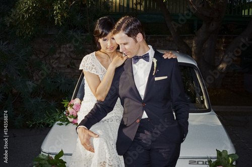 Man and woman sitting on car bonnet wearing dress and suit 