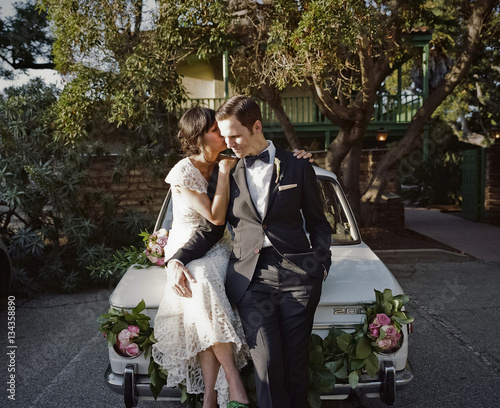 Man and woman sitting on car bonnet wearing dress and suit 