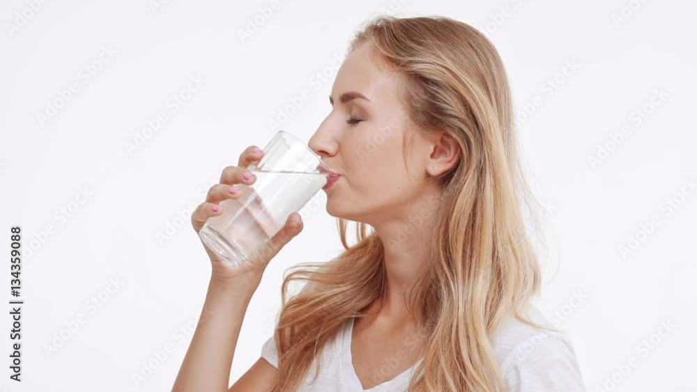 Young beautiful Caucasian blonde girl drinking water from glass and smiling at camera. Profile view on white background in slowmotion Young beautiful Caucasian blonde girl drinking water from glass and smiling at camera. Profile view on white background in slowmotion