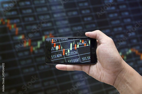 A hand holding a black smartphone showing stock market candlestick trend on blurred-out stock market data display panel
