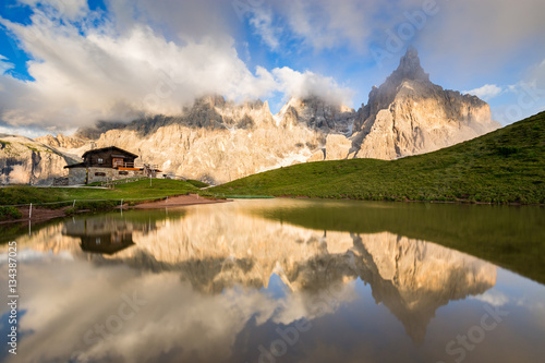 The Pale di San Martino peaks (Italian Dolomites) reflected in the water, with an alpine chalet on background.