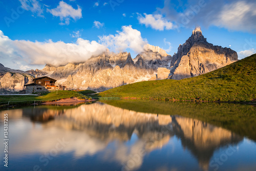 The Pale di San Martino peaks (Italian Dolomites) reflected in the water at sunset, with an alpine chalet on background.
