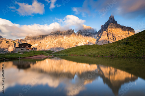 The Pale di San Martino peaks (Italian Dolomites) reflected in the water at sunset, with an alpine chalet on background. photo