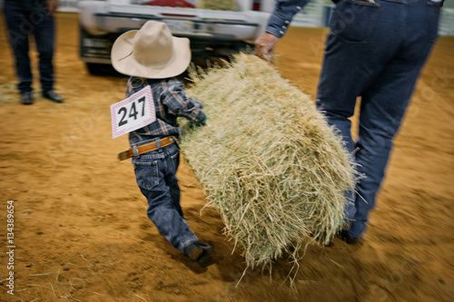 Little boy carrying a large bale of hay at a rodeo competition.