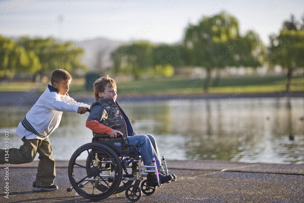 Boy pushing friend in wheelchair through park Stock Photo | Adobe Stock