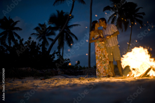 Mid-adult couple standing beside a bonfire on a beach.