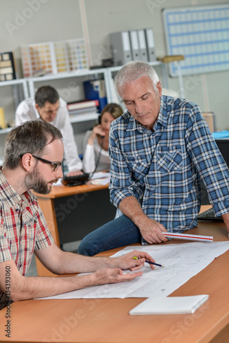 Two men looking at blueprints, one sat on the table