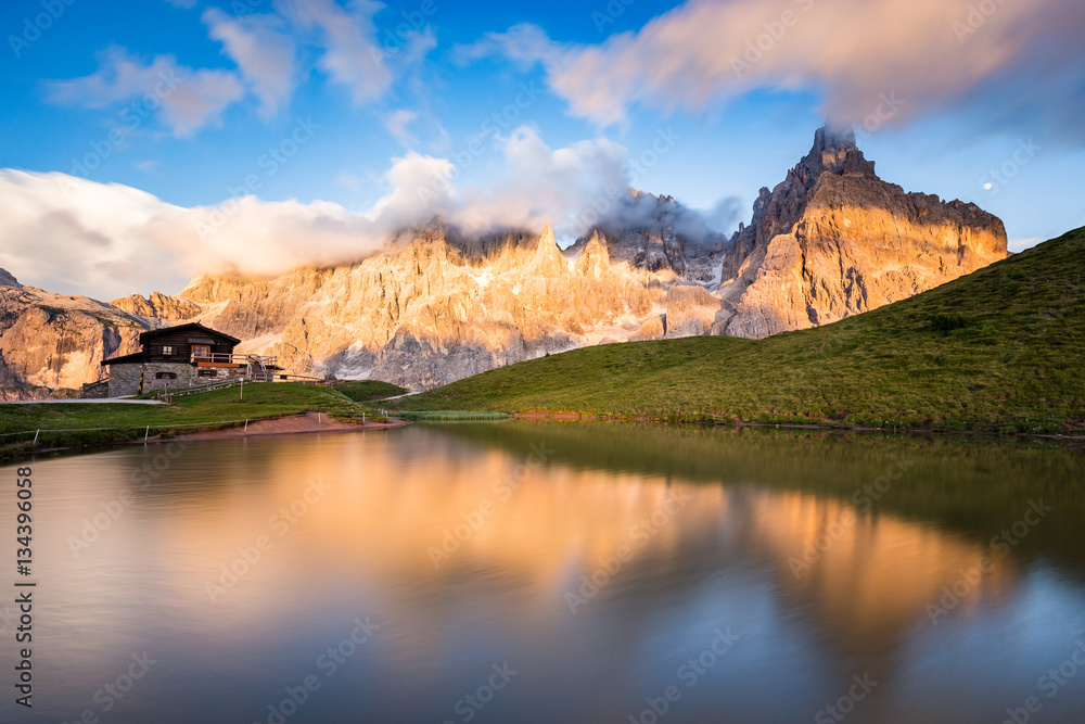 The Pale di San Martino peaks (Italian Dolomites) reflected in the water at sunset, with an alpine chalet on background.