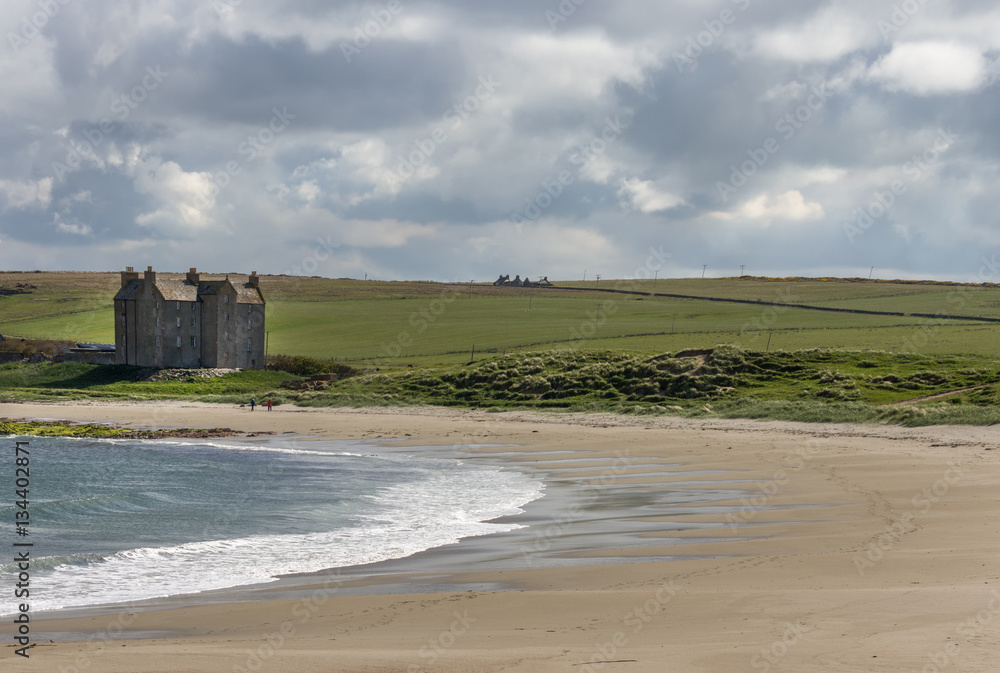 Freswidk Bay, Scotland - June 4, 2012: Wide shot of the strong house or castle, now a large farm, standing right at the beach of the bay. Cloudy sky above green meadows. Sandy beach and North Sea.