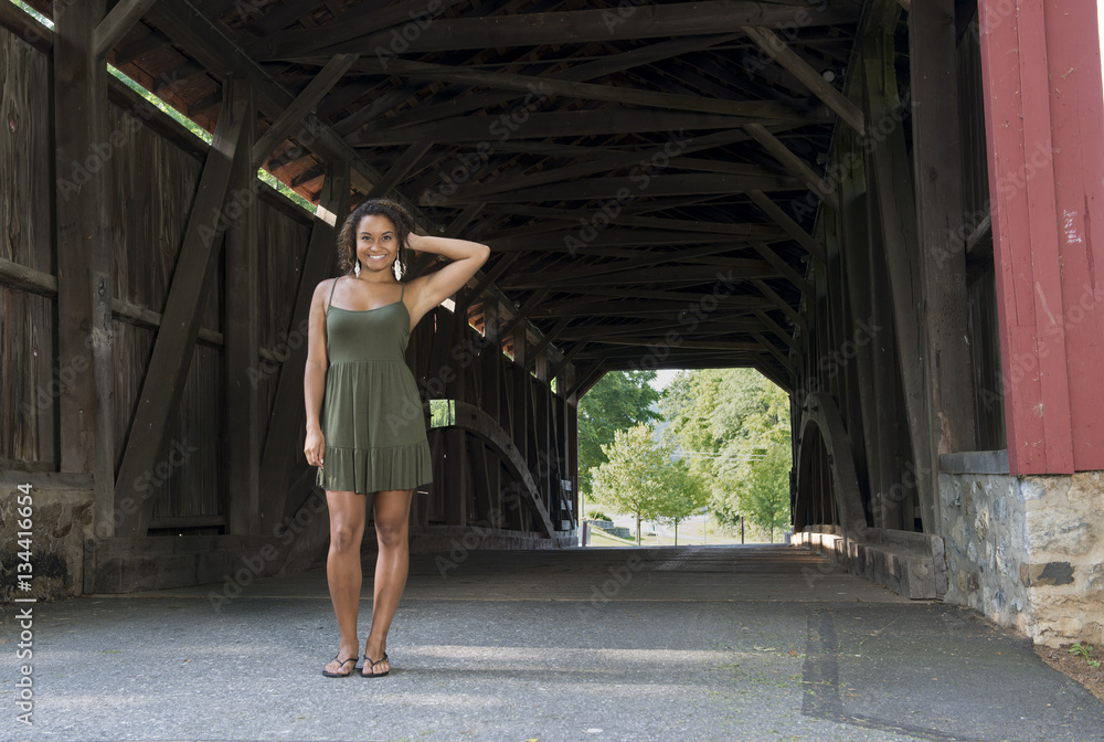 Naklejka premium Stunning young biracial woman in green sundress standing in front of covered bridge