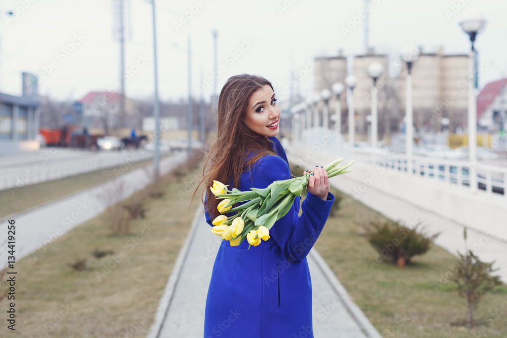 Young attractive girl in a blue coat and yellow dress holding a bouquet of yellow tulips. Spring is coming to town. She walks down the sidewalk and turned back