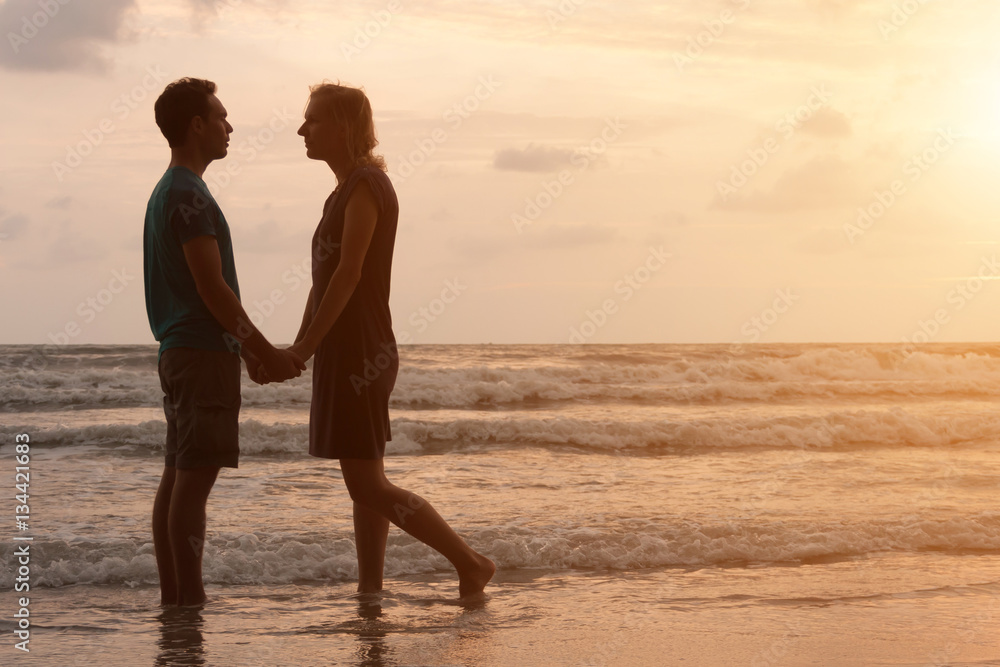 Couple in love is staying on beautiful sunset beach, holding their hands and looking on each other. Love and romance in vacation honeymoon.