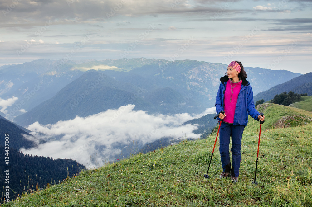Young woman with trekking poles at mountain trail. View from above on a valley with fog and clouds