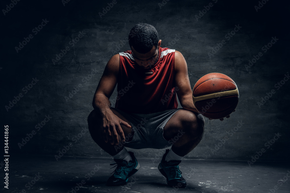 A black basketball player sits on a floor and holds basket ball.