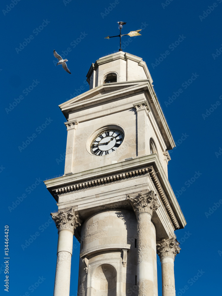 The newly cleaned and refurbished Herne Bay clock tower, in Kent, Uk with a seagull flying by on a summer day with a blue sky .