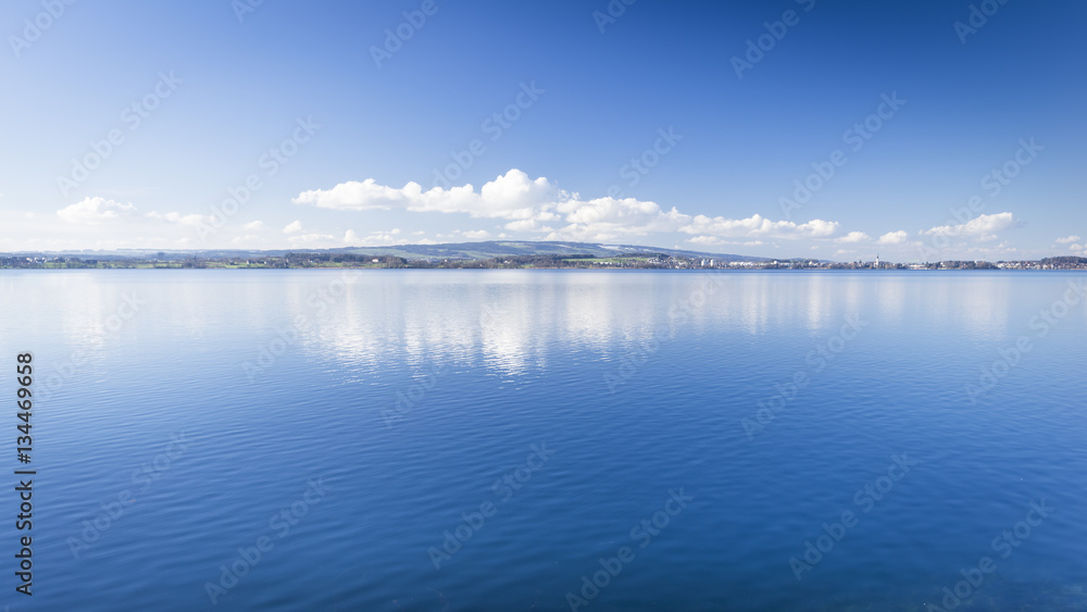 Fototapeta premium Clear day. Lake Zug in central Switzerland. Clouds are reflected in water lake. At the edge water is clear and see the bottom of the lake.