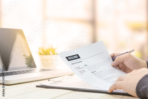 Businessman review his resume on his wooden desk before sending to find a job with laptop computer, clock and glasses.