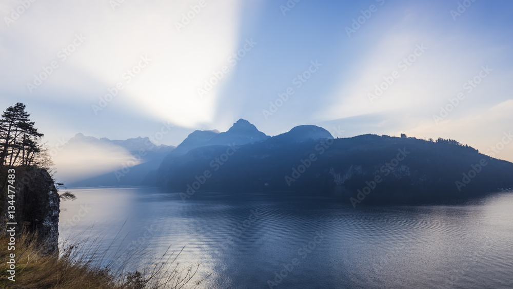 Alps of central Switzerland. Grand game of light and shadow. Sunlight sat behind a mountain ridge. Rays of light pierce the sky. On water of the lake waves and light reflection .