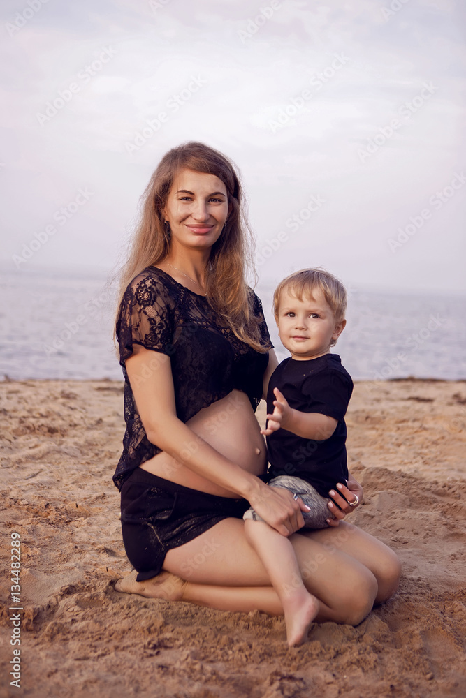 young pregnant woman in black clothes on the shore of the sea at sunset sitting with his son on the knees