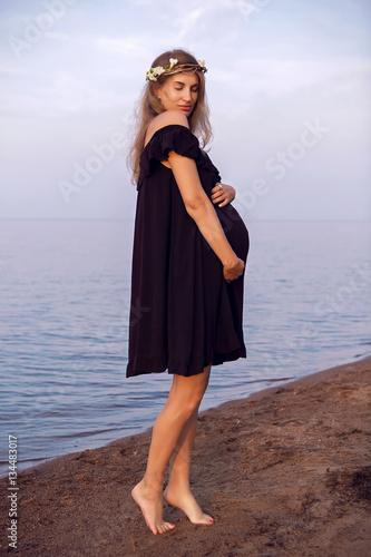 pregnant woman with long hair standing on the beach in a black dress at sunset