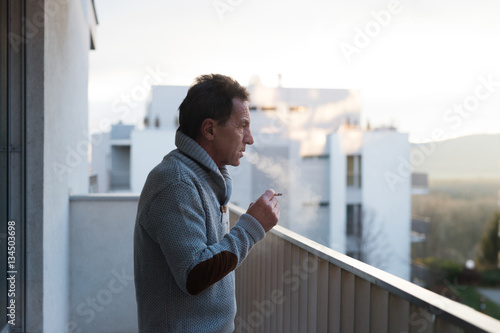 Serious senior man standing on balcony, smoking a cigarette