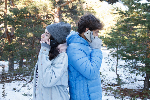 Boyfriend and girlfriend couple are talking on the phone while they are on a date in the mountains on a snowy day. They are bored and pass from each other. 