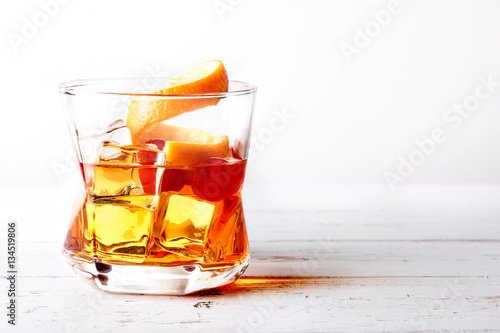 Glass of old fashioned whiskey with ice, orange and cherries over a white wood table