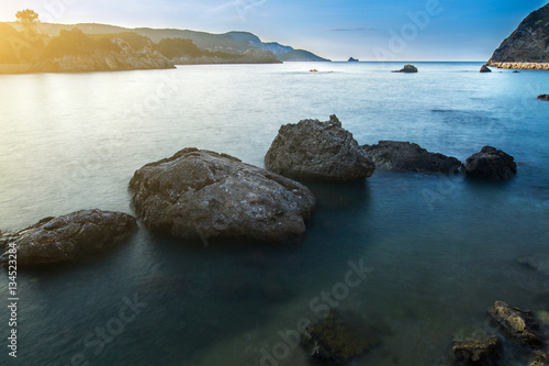 Long exposure seascape at sunrise. View of the cliff into the sea and distant islands. In the backlight sunbeam light. Paleokastrica. Corfu. Ionian archipelago. Greece.