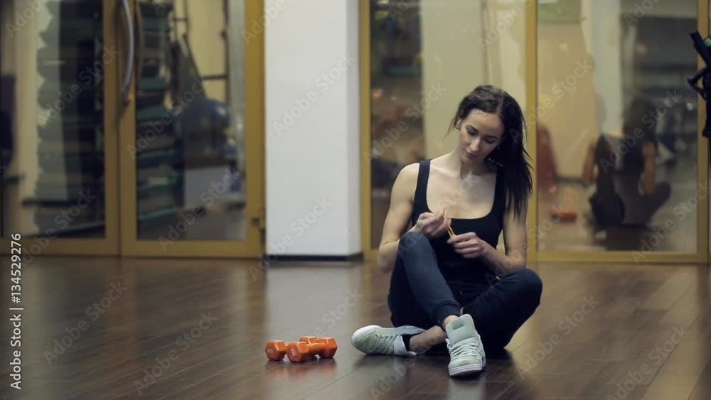In gym after exercise with gantellyami woman corrects her hair. Young slim sportswoman sitting on the floor and resting after a workout with orange weighting, which lie near legs. During a break, she In gym after exercise with gantellyami woman corrects her hair. Young slim sportswoman sitting on the floor and resting after a workout with orange weighting, which lie near legs. During a break, she