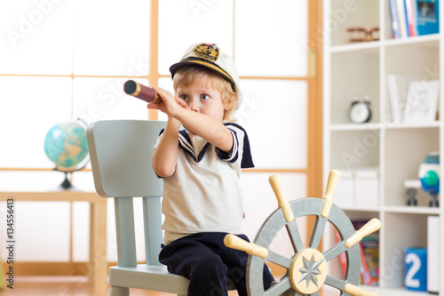 Kid boy dressed like a captain or sailor plays on chair as ship in his room. Child looks through telescope. photo