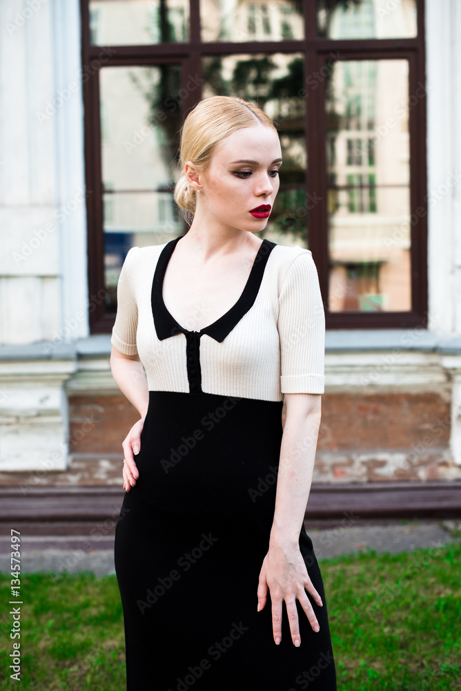 Happy Fashion blonde european elegant Woman with red lips and white skin Standing at the Old Red Brick building on green grass. Beautiful Female in formal black and white dress. Happy Fashion blonde european elegant Woman with red lips and white skin Standing at the Old Red Brick building on green grass. Beautiful Female in formal black and white dress.